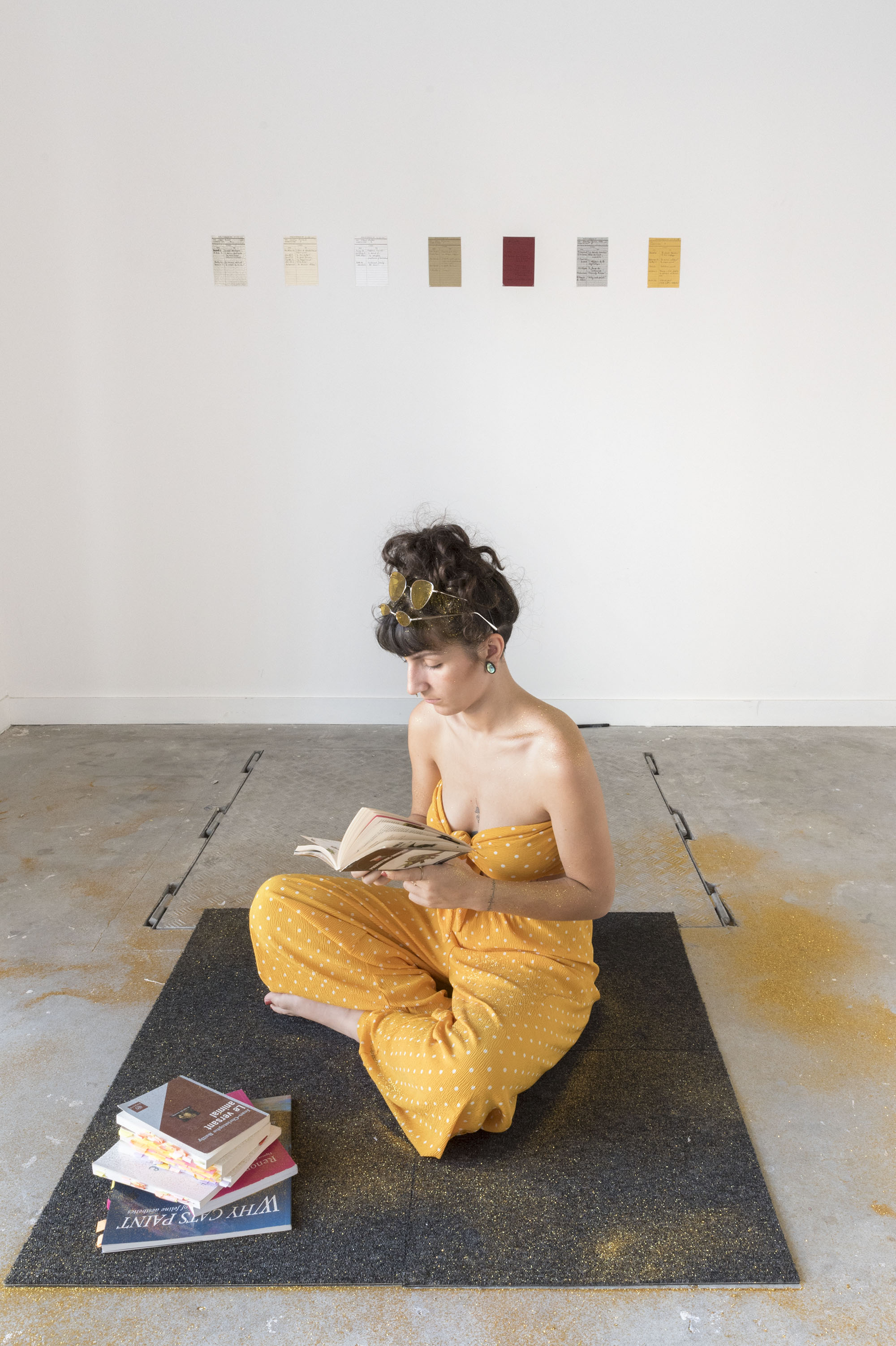 Une jeune femme lit, assise sur un carré de moquette. Une pile de livres est posé à côté d'elle. Au mur on voit 7 petites fiches accrochées.